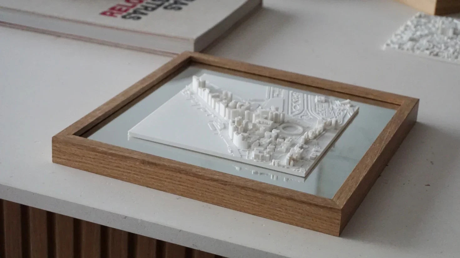 Framed 3D city map in white, with raised buildings, displayed on a modern tabletop