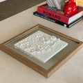 Framed 3D city map decor in white, displayed on a table beside stacked books.